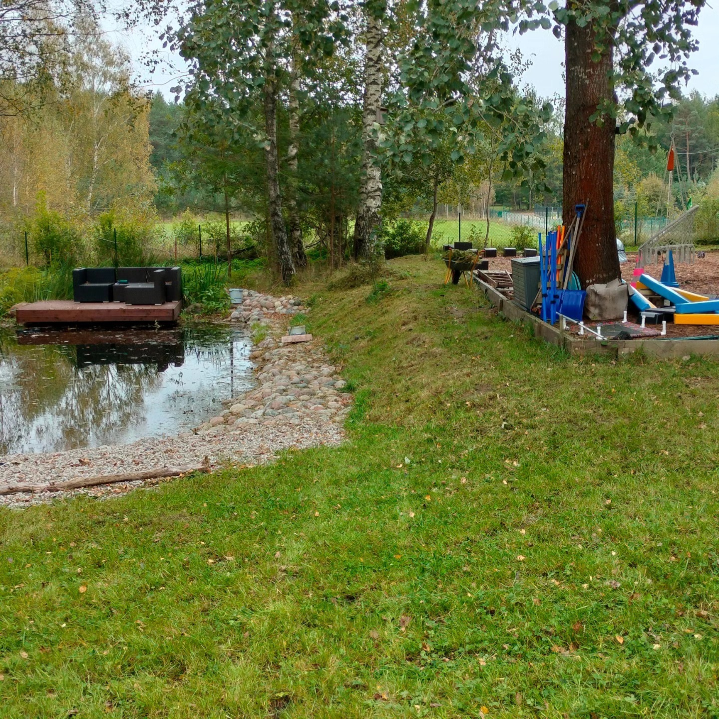 Widok na oczko wodne z drewnianym pomostem, na którym stoi czarny, nowoczesny zestaw mebli ogrodowych; brzeg oczka wyłożony kamieniami, w tle drzewa i trawnik z narzędziami ogrodniczymi opartymi...