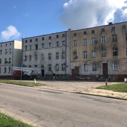 Profesjonalne czyszczenie elewacji budynków, dachów oraz kostki brukowej, Mateusz Leniar - Wielopiętrowy budynek mieszkalny z brudną elewacją, częściowo wyczyszczoną, z widocznym podnośnikiem koszowym i pracownikiem na ziemi, sugerującym prace związane z myciem elewacji.