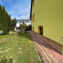 Żółta elewacja budynku z dekoracyjnymi obramowaniami okien, widoczna z perspektywy trawnika z pomarańczowym wężem ogrodowym i ścieżką z kostki brukowej w dwóch kolorach.