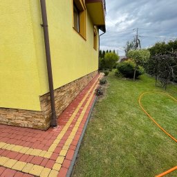 Profesjonalne czyszczenie elewacji budynków, dachów oraz kostki brukowej, Mateusz Leniar - Żółta elewacja domu z kamienną podmurówką i chodnikiem z czerwono-żółtej kostki brukowej, obok zielony trawnik z pomarańczowym wężem ogrodowym w pochmurny dzień.