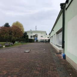 Profesjonalne czyszczenie elewacji budynków, dachów oraz kostki brukowej, Mateusz Leniar - Okrągła myjka ciśnieniowa na kostce brukowej, widoczne budynki w tle, jesienne drzewa i ławki.