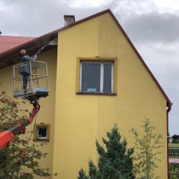 Profesjonalne czyszczenie elewacji budynków, dachów oraz kostki brukowej, Mateusz Leniar - Pracownik na podnośniku koszowym czyści żółtą elewację domu za pomocą myjki ciśnieniowej, widoczna różnica między umytą a brudną częścią ściany.
