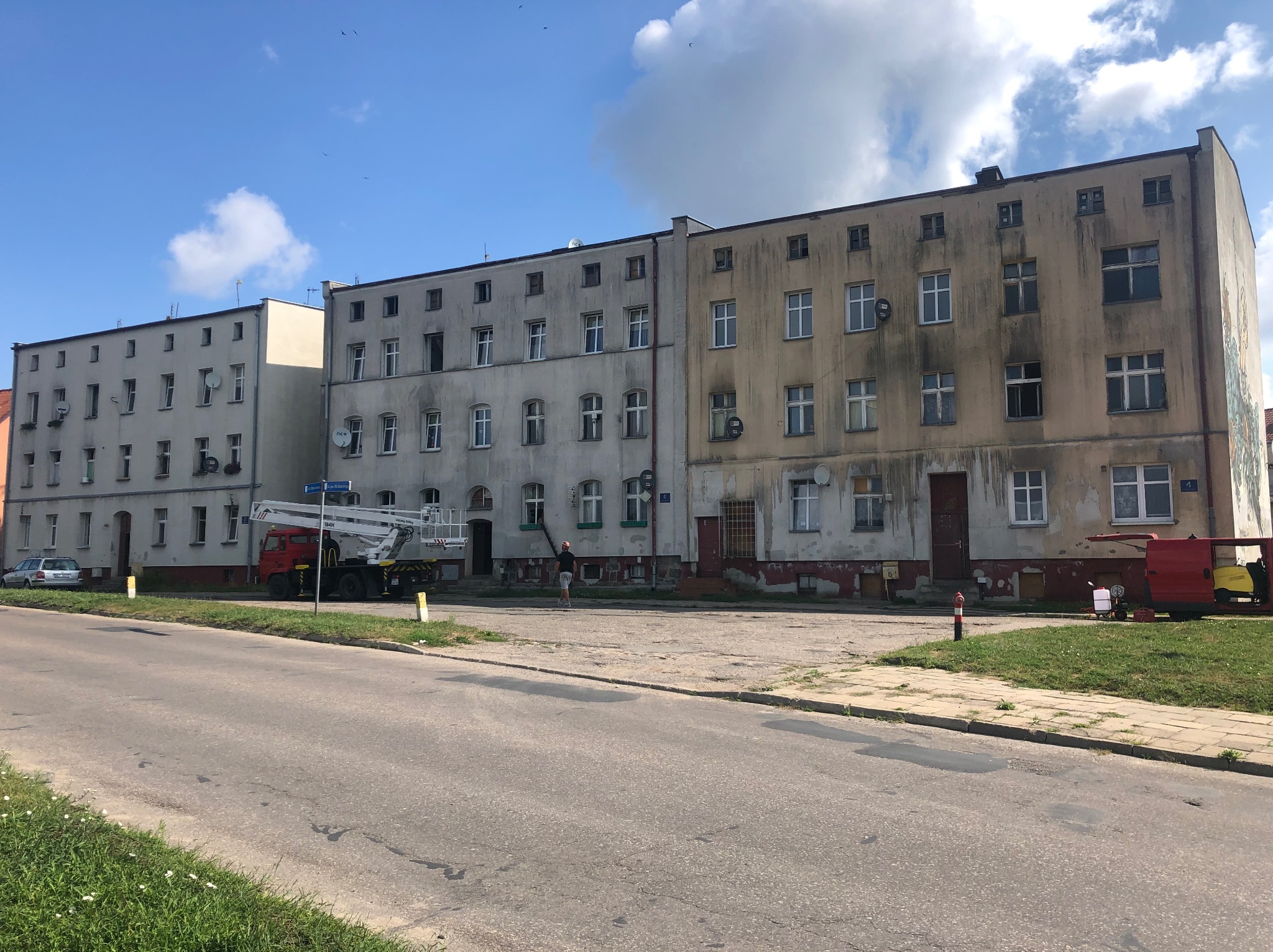 Wielopiętrowy budynek mieszkalny z brudną elewacją, częściowo wyczyszczoną, z widocznym podnośnikiem koszowym i pracownikiem na ziemi, sugerującym prace związane z myciem elewacji.