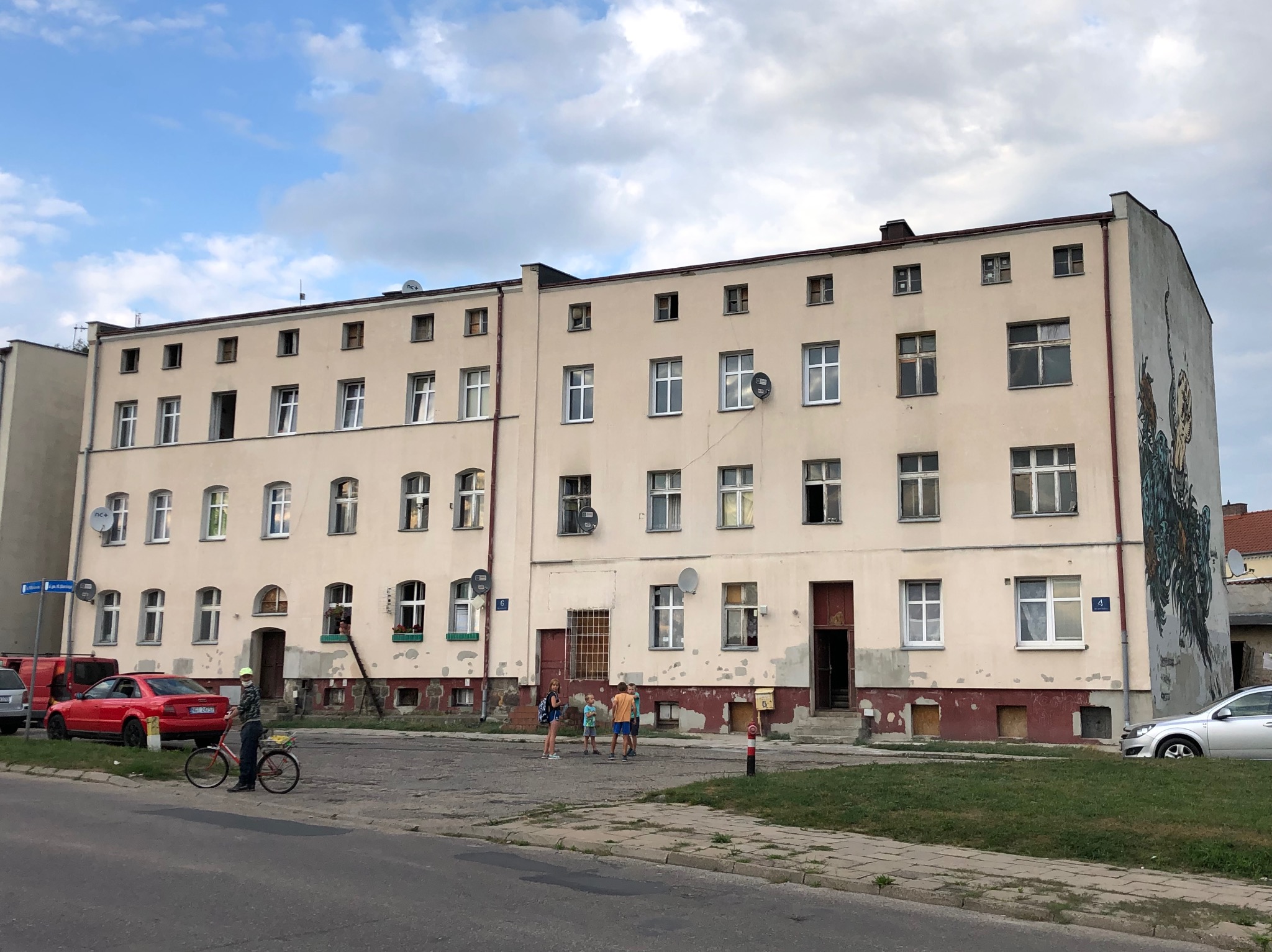 Trzy piętrowy budynek mieszkalny z widocznymi śladami zabrudzeń i odchodzącej farby na elewacji, z muralem na ścianie szczytowej.