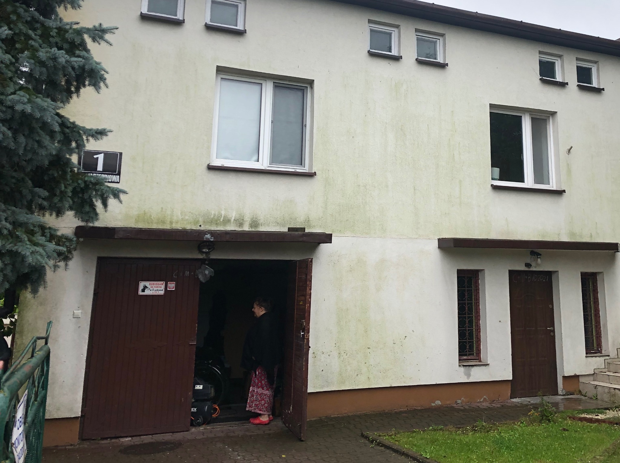 Dwukondygnacyjny budynek z garażem i widocznymi zabrudzeniami na elewacji, z otwartych drzwi garażowych wychodzi kobieta.
