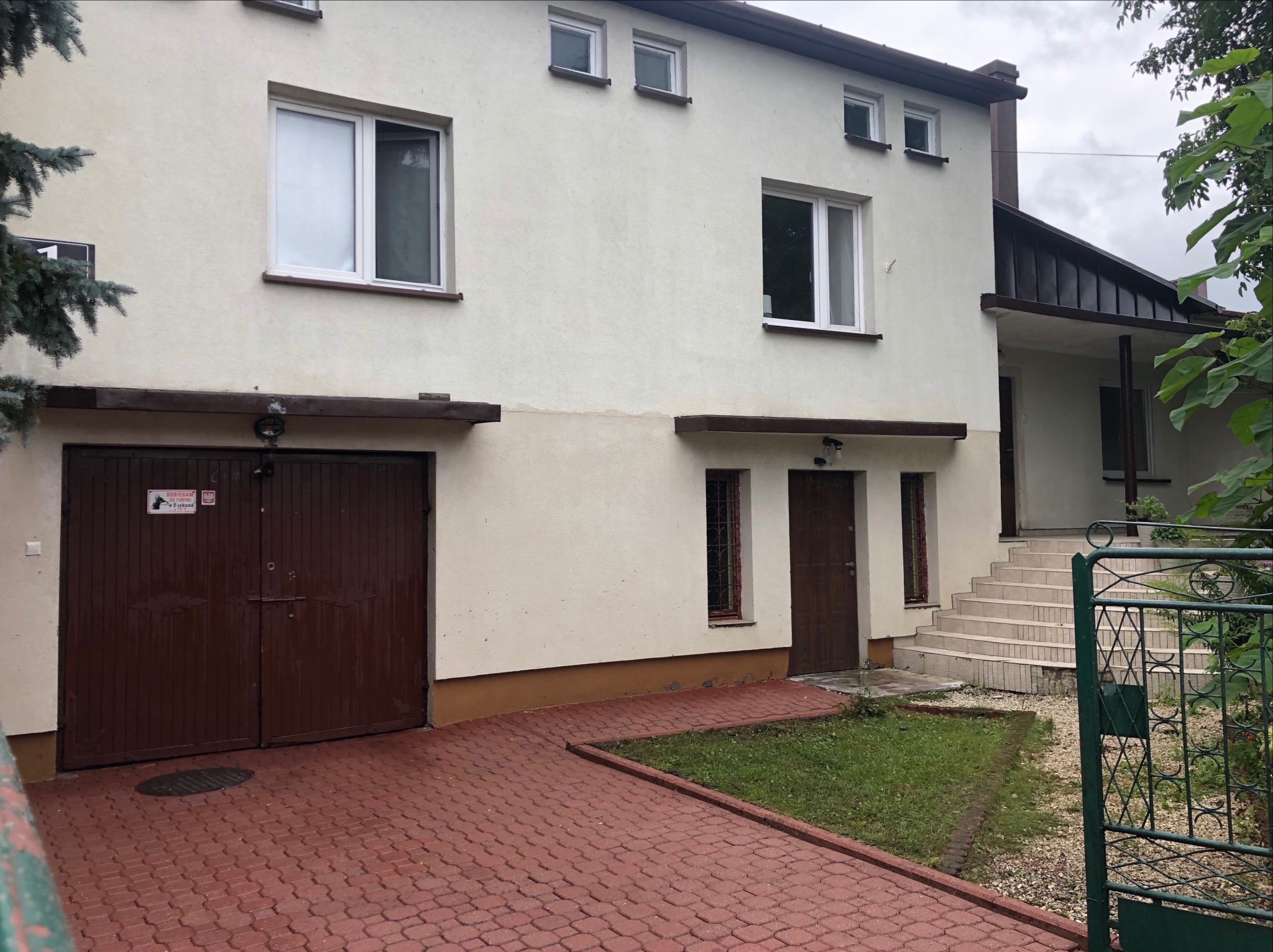 Dwupiętrowy dom z garażem i czerwonym brukiem, widoczne okna i drzwi wejściowe, zieleń w tle, pochmurne niebo.