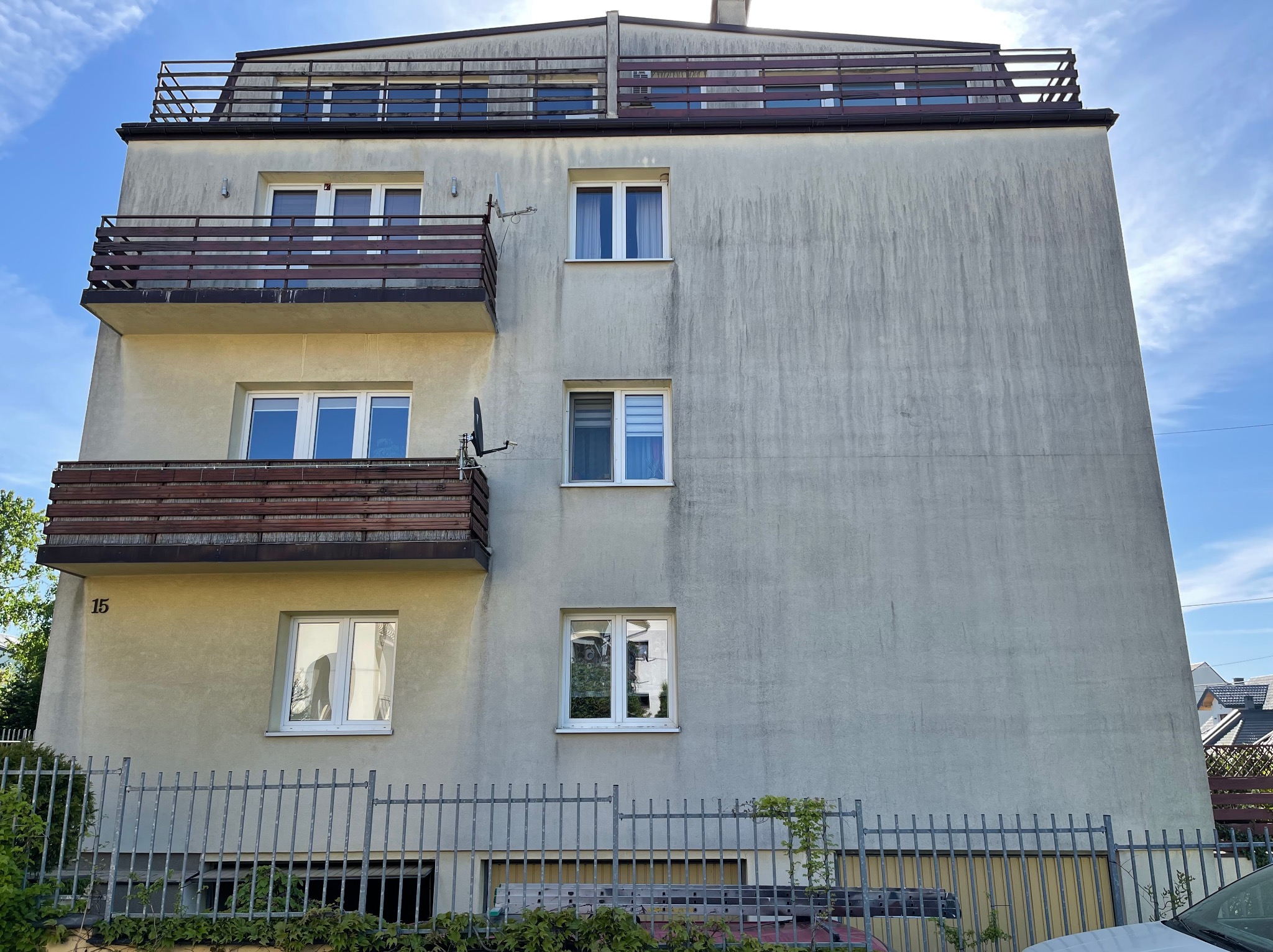 Wielopiętrowy budynek z jasną elewacją wymagającą odświeżenia, widoczne zacieki i zabrudzenia, balkony z drewnianymi barierkami, numer 15 na ścianie przy gruncie.