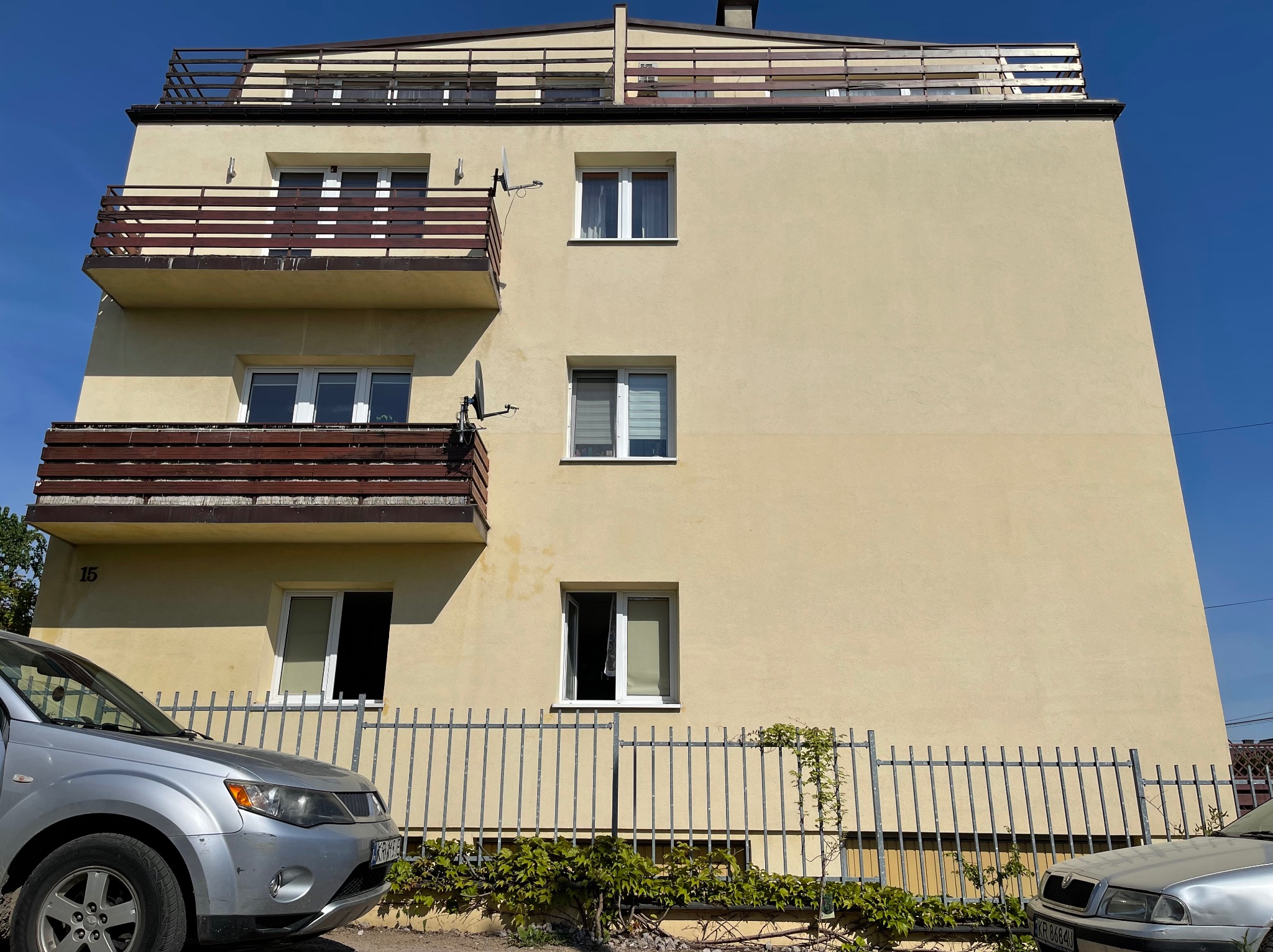 Żółta elewacja budynku z trzema balkonami, widoczne samochody i metalowe ogrodzenie z pnącą roślinnością u dołu