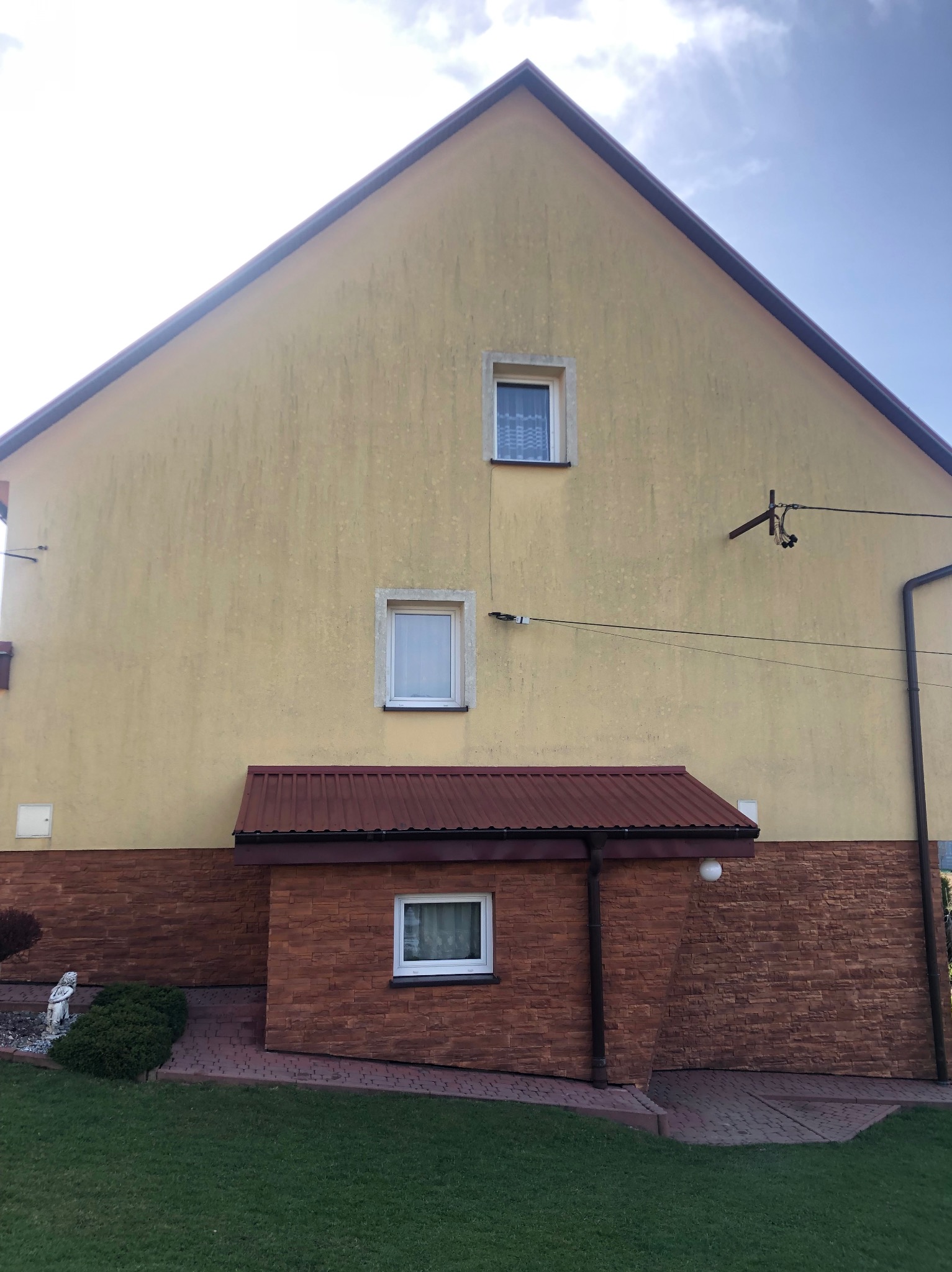 Dwupiętrowy dom z żółtą elewacją, widoczne ślady zabrudzeń i zacieków, brązowa podmurówka z cegły, trzy okna z białymi ramami, trawnik i dekoracyjny krzew przed budynkiem.