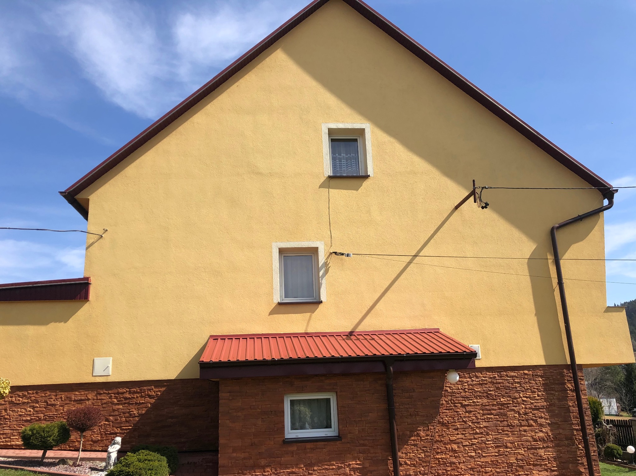 Żółta elewacja domu z brązowym dachem i kamienną dolną częścią, widoczne dwa małe okna na górze i jedno większe na dole, błękitne niebo w tle.