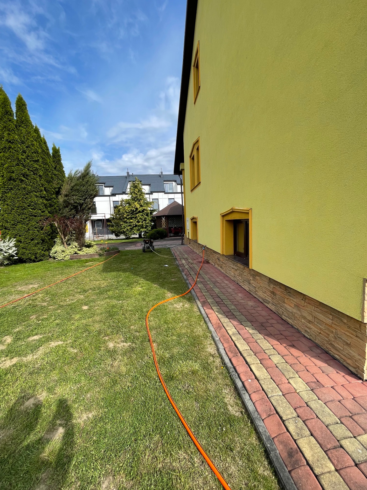 Żółta elewacja budynku z dekoracyjnymi obramowaniami okien, widoczna z perspektywy trawnika z pomarańczowym wężem ogrodowym i ścieżką z kostki brukowej w dwóch kolorach.