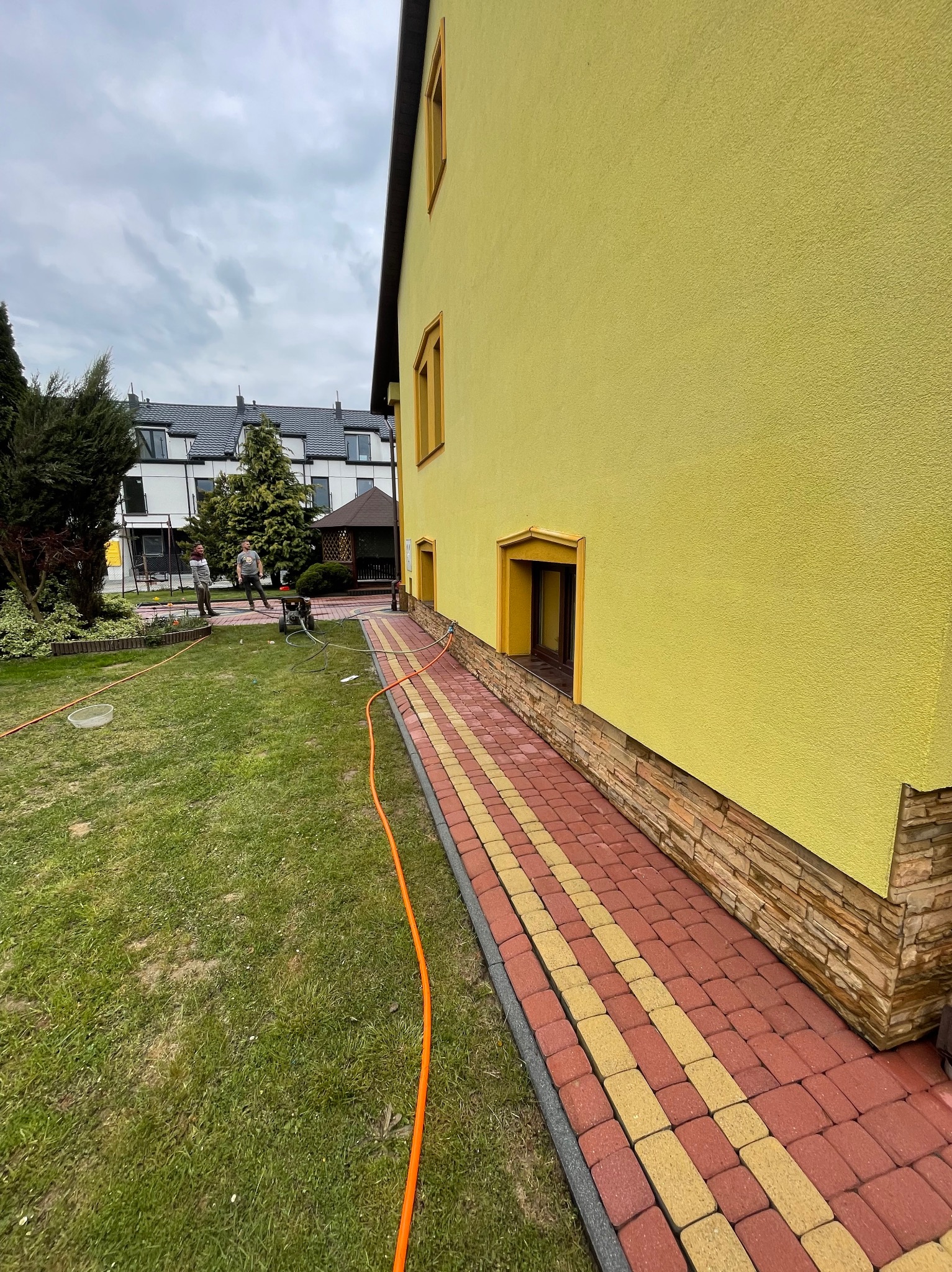 Żółta elewacja domu w Rzeszowie z widoczną kamienną podmurówką i chodnikiem z kostki brukowej, przygotowana do czyszczenia. W tle pracownicy z myjką ciśnieniową.