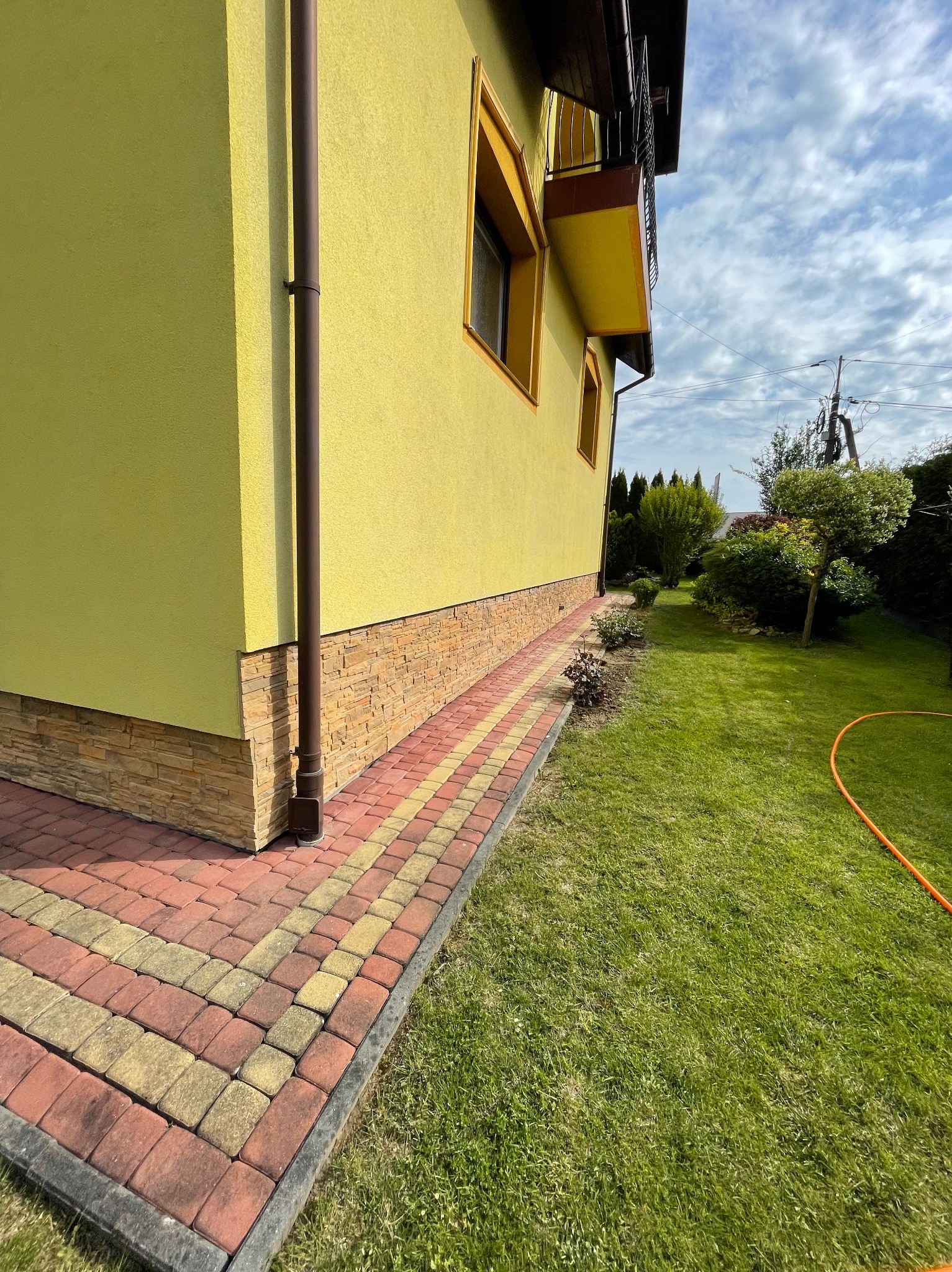 Żółta elewacja domu z dekoracyjnym kamieniem u podstawy, widoczny fragment rynny spustowej i kolorowa kostka brukowa na obrzeżach trawnika.