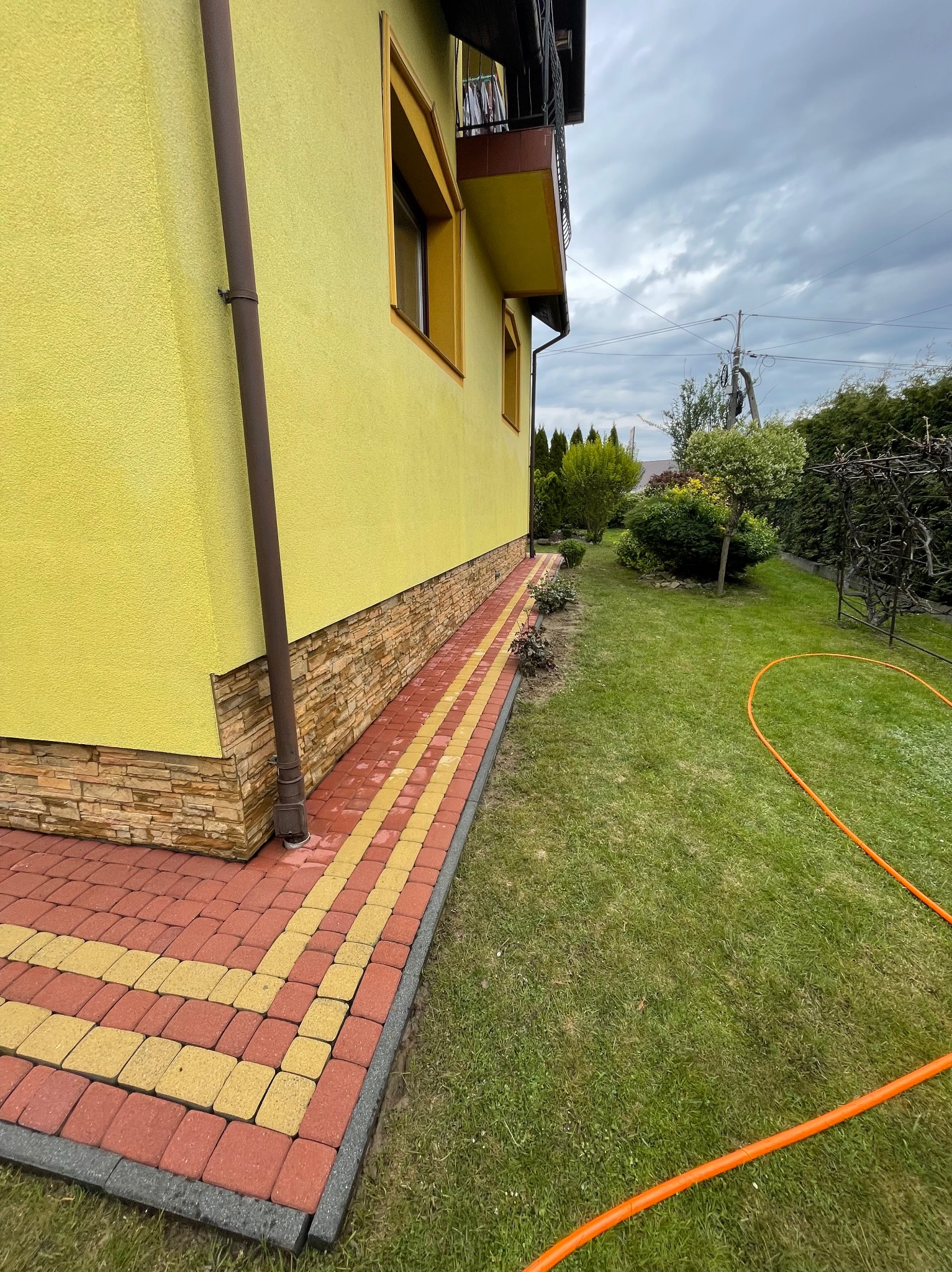 Żółta elewacja domu z kamienną podmurówką i chodnikiem z czerwono-żółtej kostki brukowej, obok zielony trawnik z pomarańczowym wężem ogrodowym w pochmurny dzień.