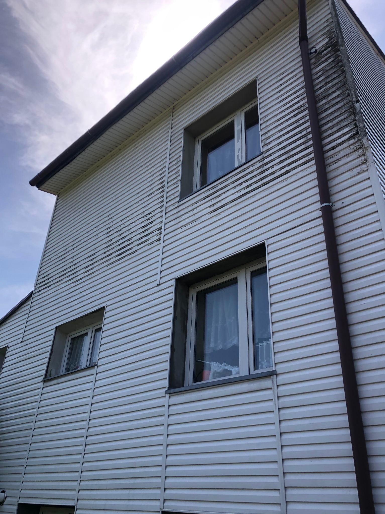 Boczna ściana domu z białym sidingiem, widoczne zabrudzenia i ślady biologiczne, brązowa rynna wzdłuż narożnika, trzy okna z białymi ramami, pochmurne niebo w tle.