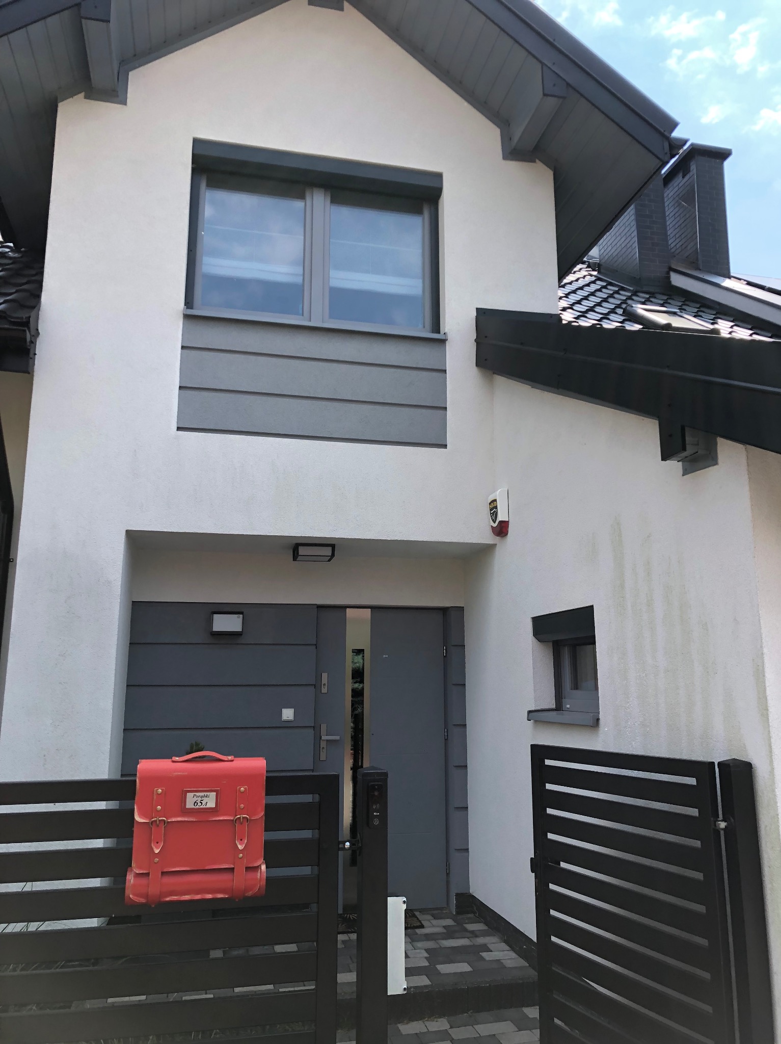 Nowoczesny dom z białą elewacją i szarymi akcentami, widoczny czerwony listonosz na ogrodzeniu, ślady zabrudzeń na ścianie obok okna.