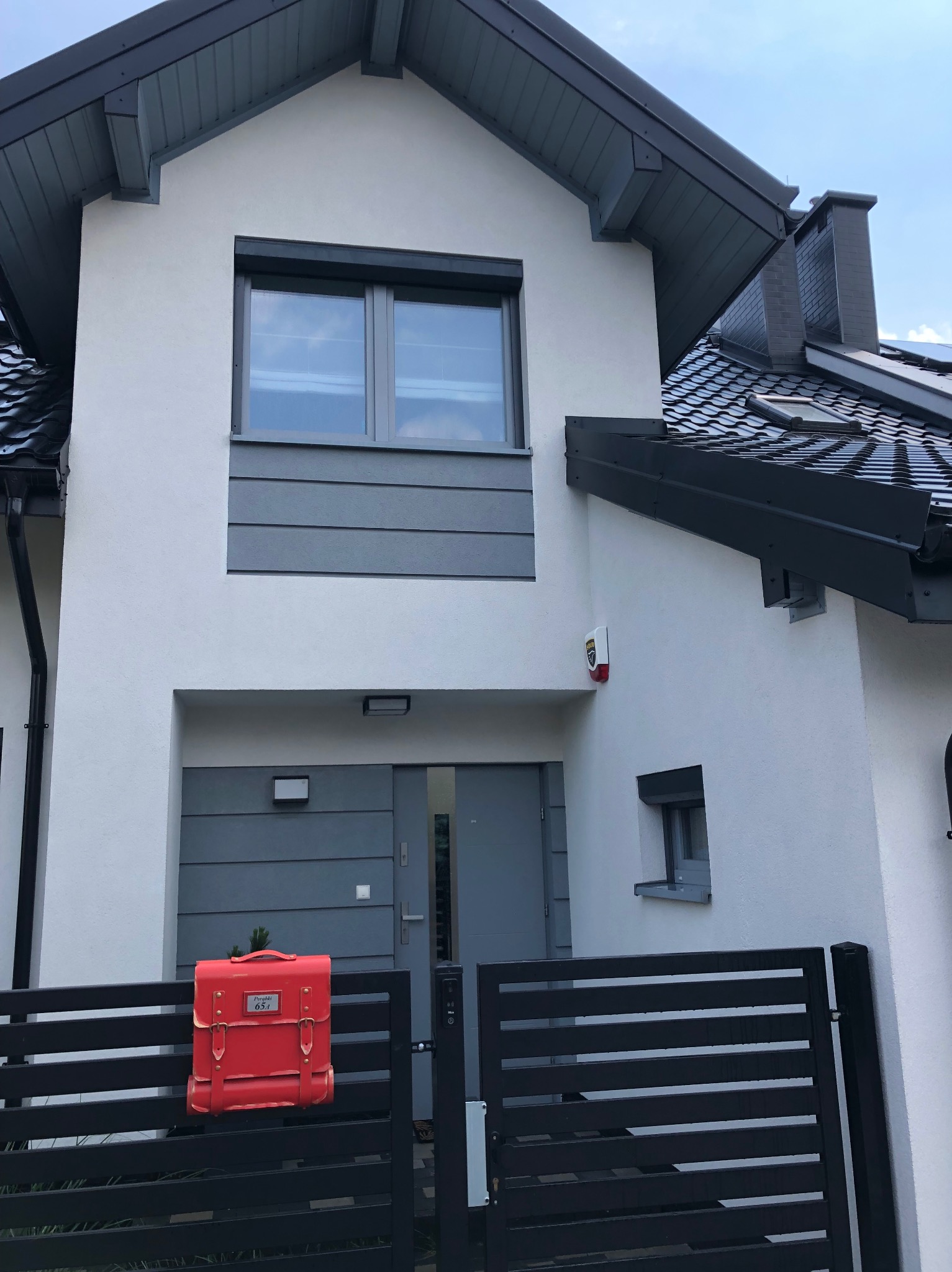 Nowoczesny dom z białą elewacją, grafitowym dachem i stolarką okienną, z czerwonym plecakiem-skrzynką pocztową na ogrodzeniu.
