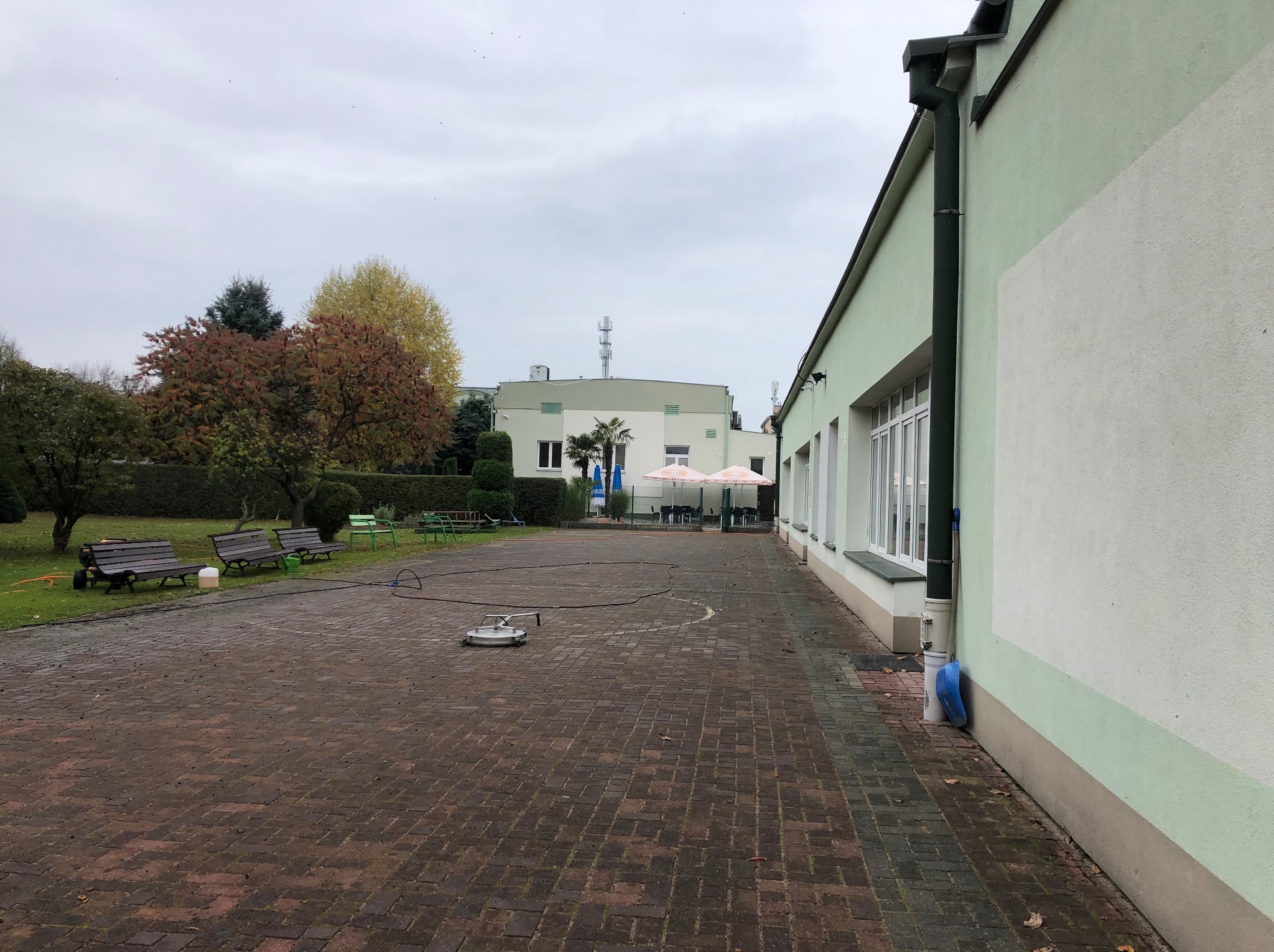 Okrągła myjka ciśnieniowa na kostce brukowej, widoczne budynki w tle, jesienne drzewa i ławki.