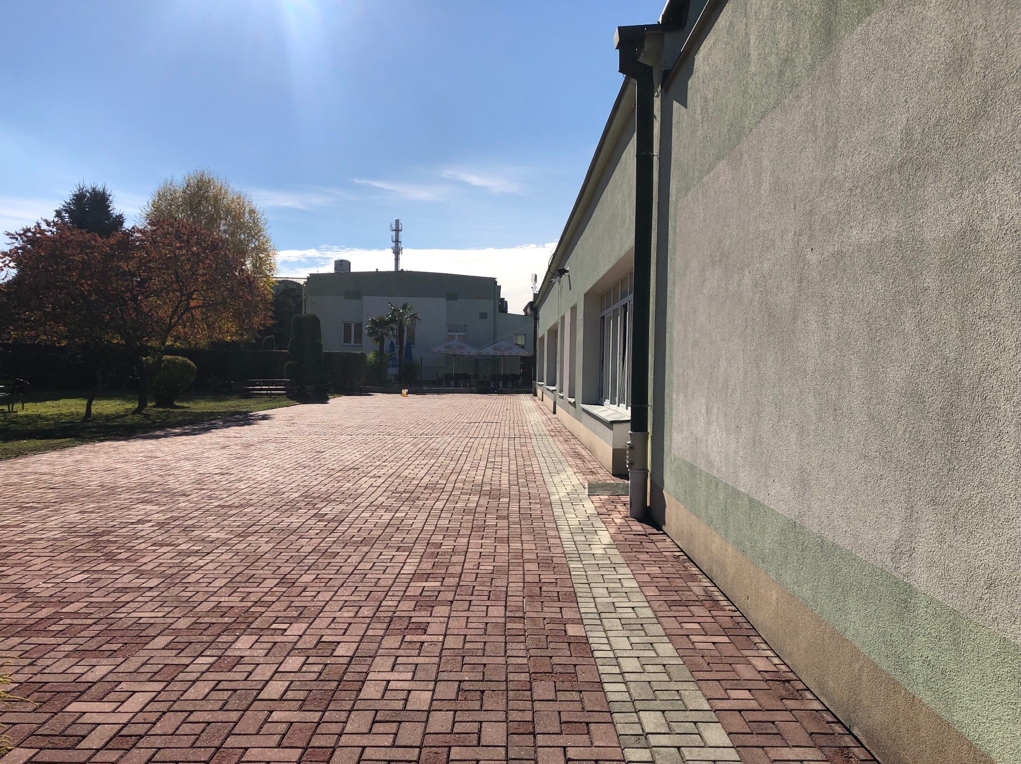 Budynek z elewacją w odcieniach szarości i zieleni, obok czerwona kostka brukowa z szarym pasem, widok z perspektywy chodnika w słoneczny dzień.