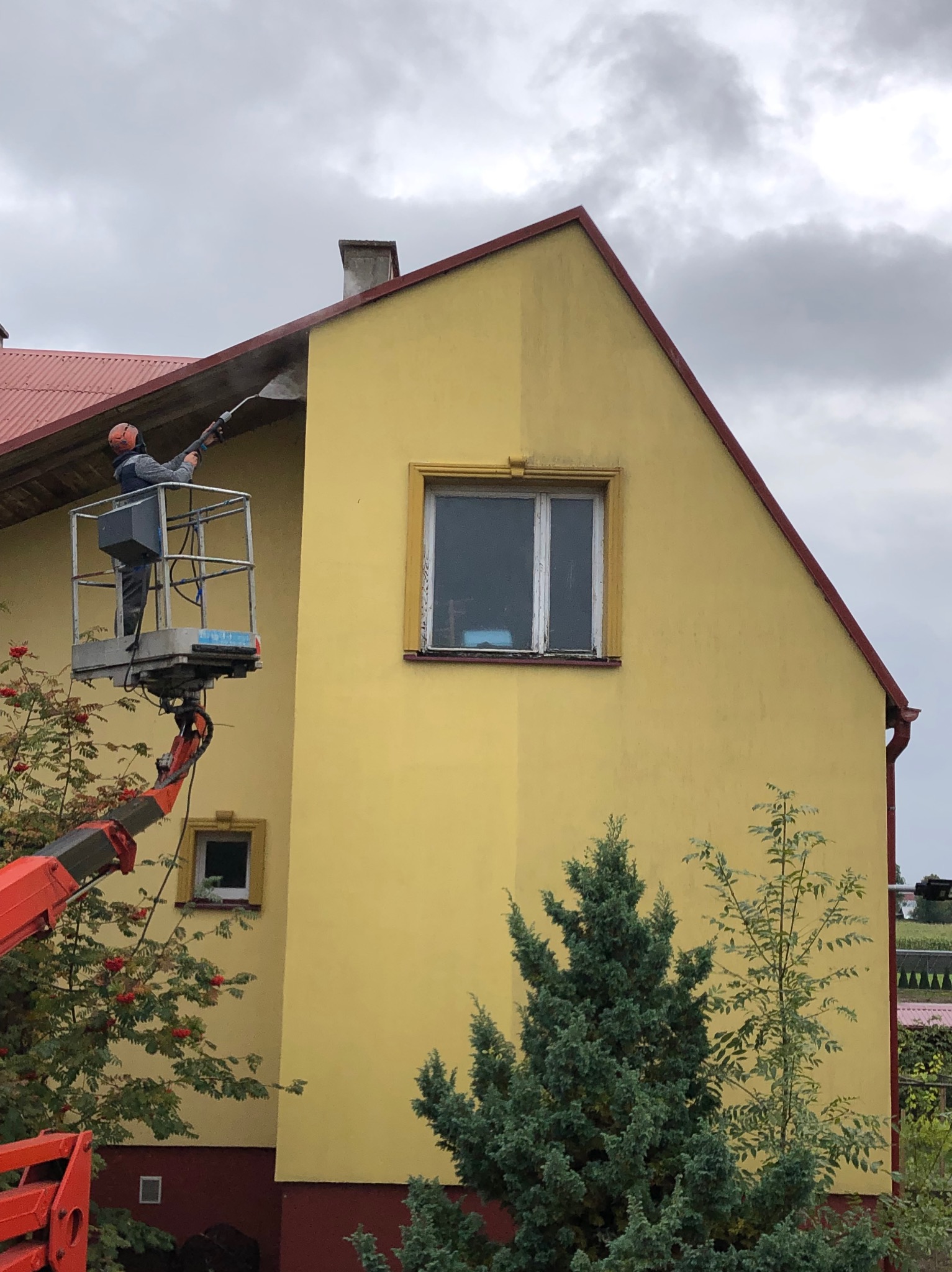 Pracownik na podnośniku koszowym czyści żółtą elewację domu za pomocą myjki ciśnieniowej, widoczna różnica między umytą a brudną częścią ściany.
