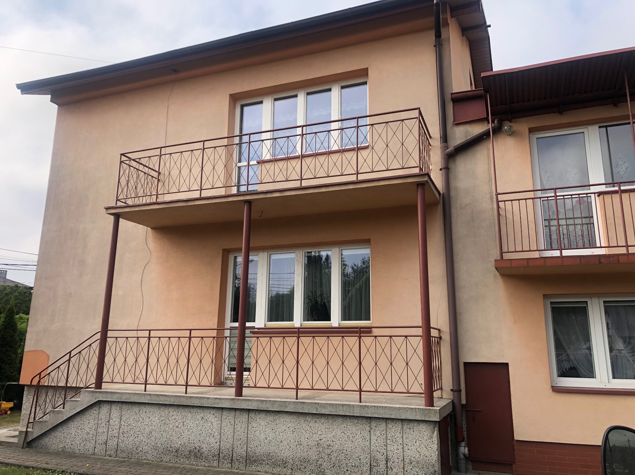 Dwupiętrowy dom z beżową elewacją i balkonami z metalowymi balustradami o geometrycznym wzorze, widoczne niewielkie pęknięcia na ścianie.