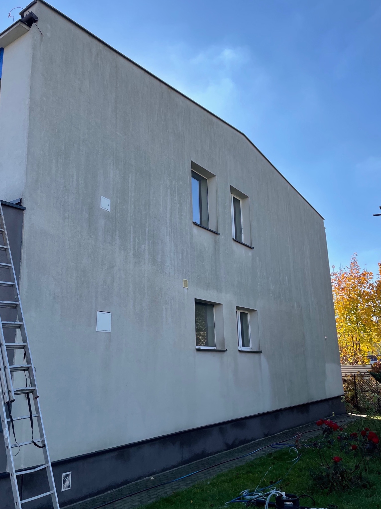 Szara elewacja budynku z widocznymi zaciekami i zabrudzeniami, z drabina opartą o ścianę i sprzętem do czyszczenia na trawniku, jesienne drzewa w tle.