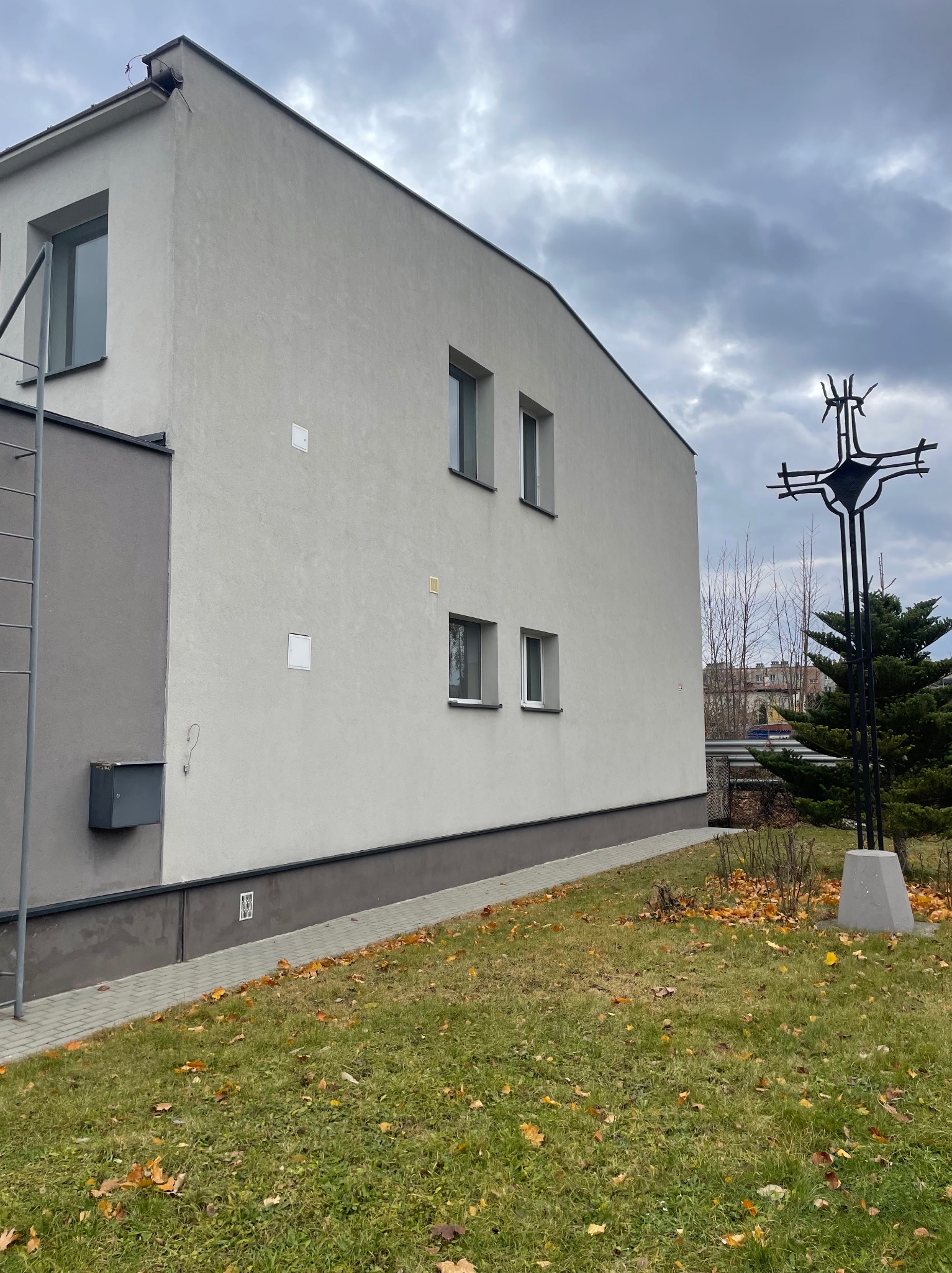 Dwukondygnacyjny budynek z jasną elewacją, widoczny z boku, z metalową konstrukcją krzyża na pierwszym planie i fragmentem chodnika z kostki brukowej.