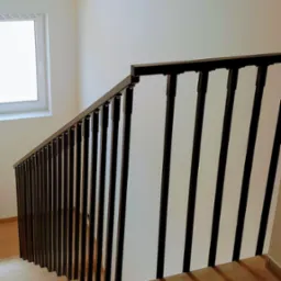 Klatka schodowa z drewnianymi schodami i czarną, minimalistyczną barierką z pionowych elementów, oświetlona przez okno.