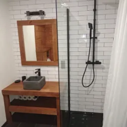 Rustykalna łazienka z drewnianą umywalką, kamienną misą, czarną armaturą i kabiną prysznicową z białymi, cegiełkowymi płytkami na ścianach.