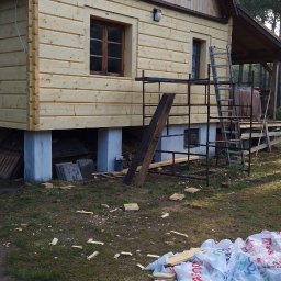 Bubla bud - Dom z drewnianą elewacją w trakcie budowy, widoczne rusztowania, okna z drewnianymi ramami, fundamenty z betonu, porozrzucane ścinki drewna i worki na trawie.