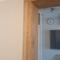 Bubla bud - Drewniana kolumna i belka stropowa z widocznym usłojeniem, stanowiące element wykończenia wnętrza, na tle jasnej ściany i fragmentu pomieszczenia z zegarem.