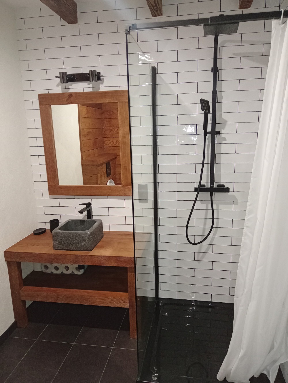 Rustykalna łazienka z drewnianą umywalką, kamienną misą, czarną armaturą i kabiną prysznicową z białymi, cegiełkowymi płytkami na ścianach.