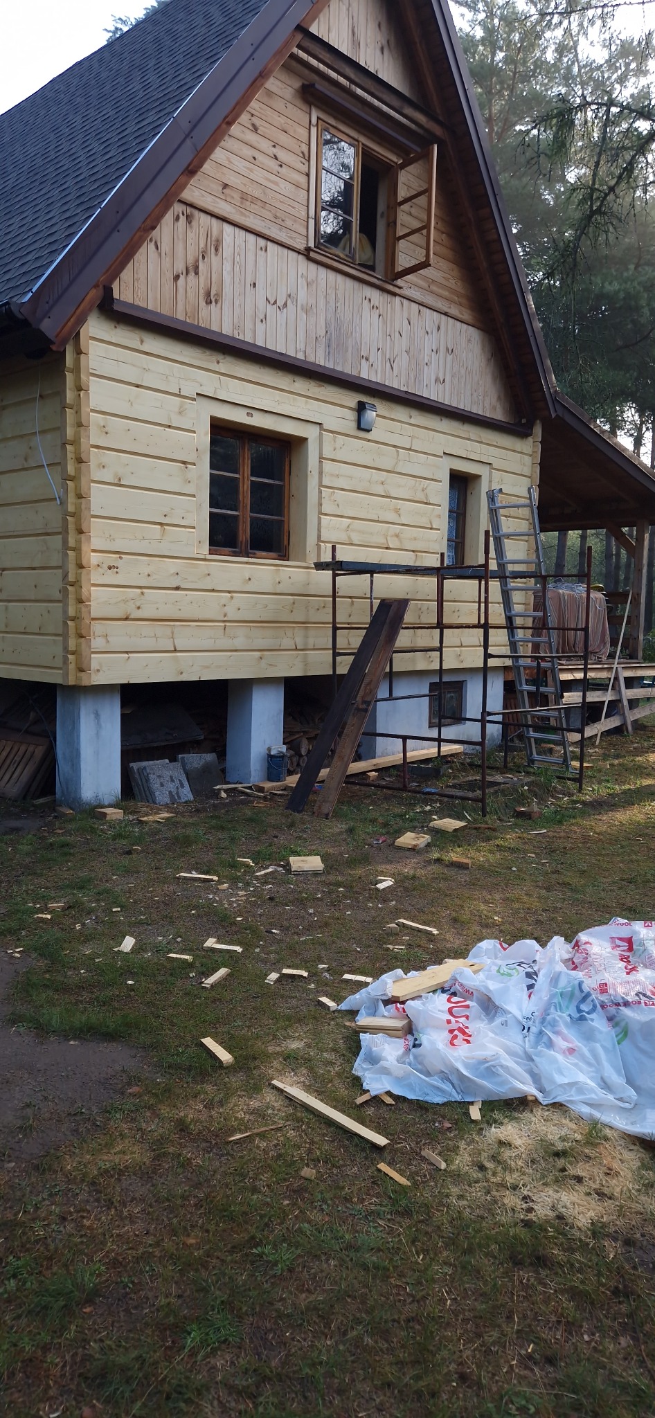 Dom z drewnianą elewacją w trakcie budowy, widoczne rusztowania, okna z drewnianymi ramami, fundamenty z betonu, porozrzucane ścinki drewna i worki na trawie.