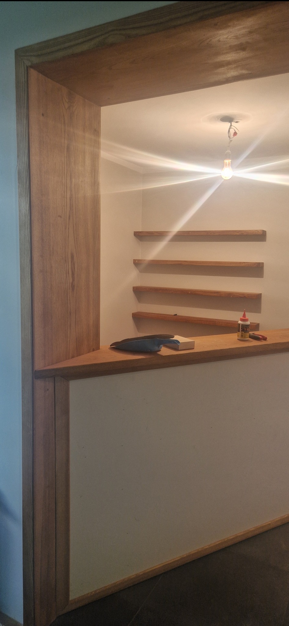 Wnęka z drewnianą ramą i półkami w trakcie montażu, widoczna żarówka bez klosza, klej i narzędzia.