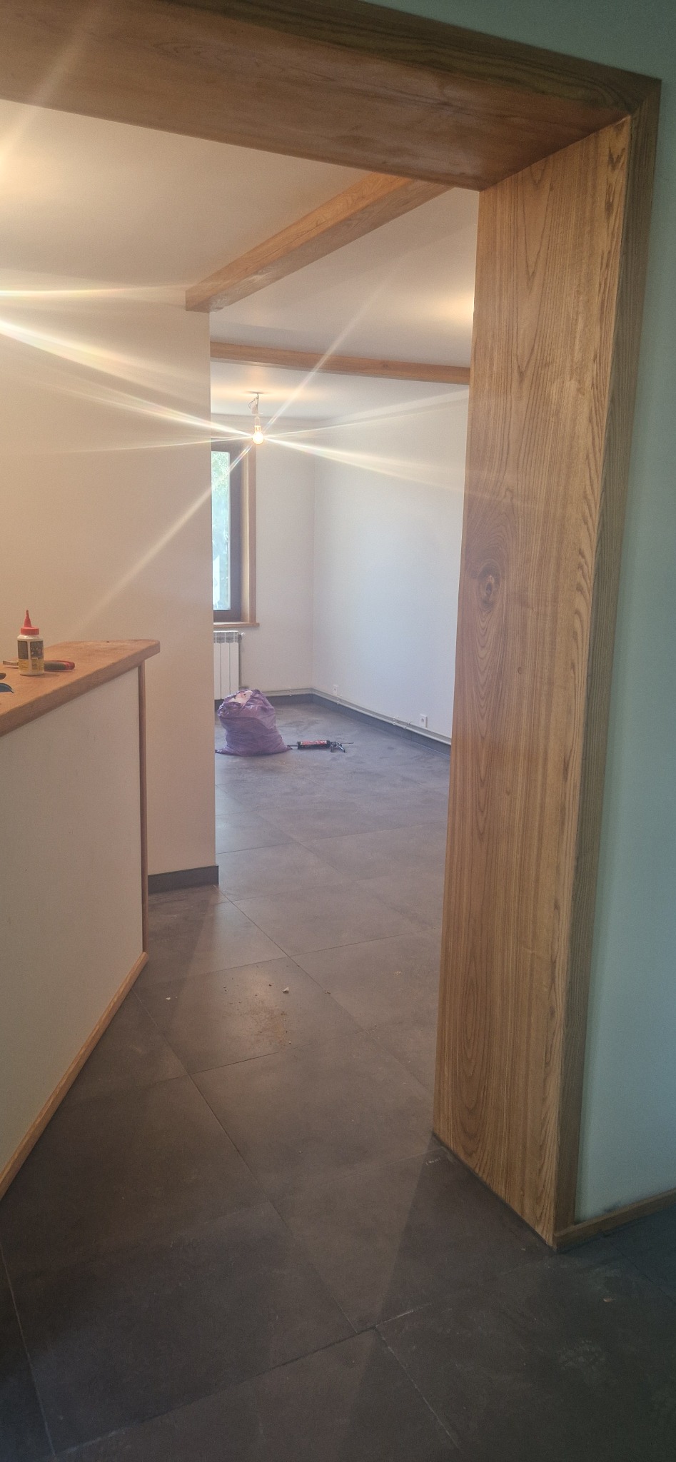 Wnętrze w trakcie remontu: widoczne drewniane wykończenia framugi i belek sufitowych, szara podłoga z płytek, ściany przygotowane do malowania, narzędzia i worek na śmieci w tle.