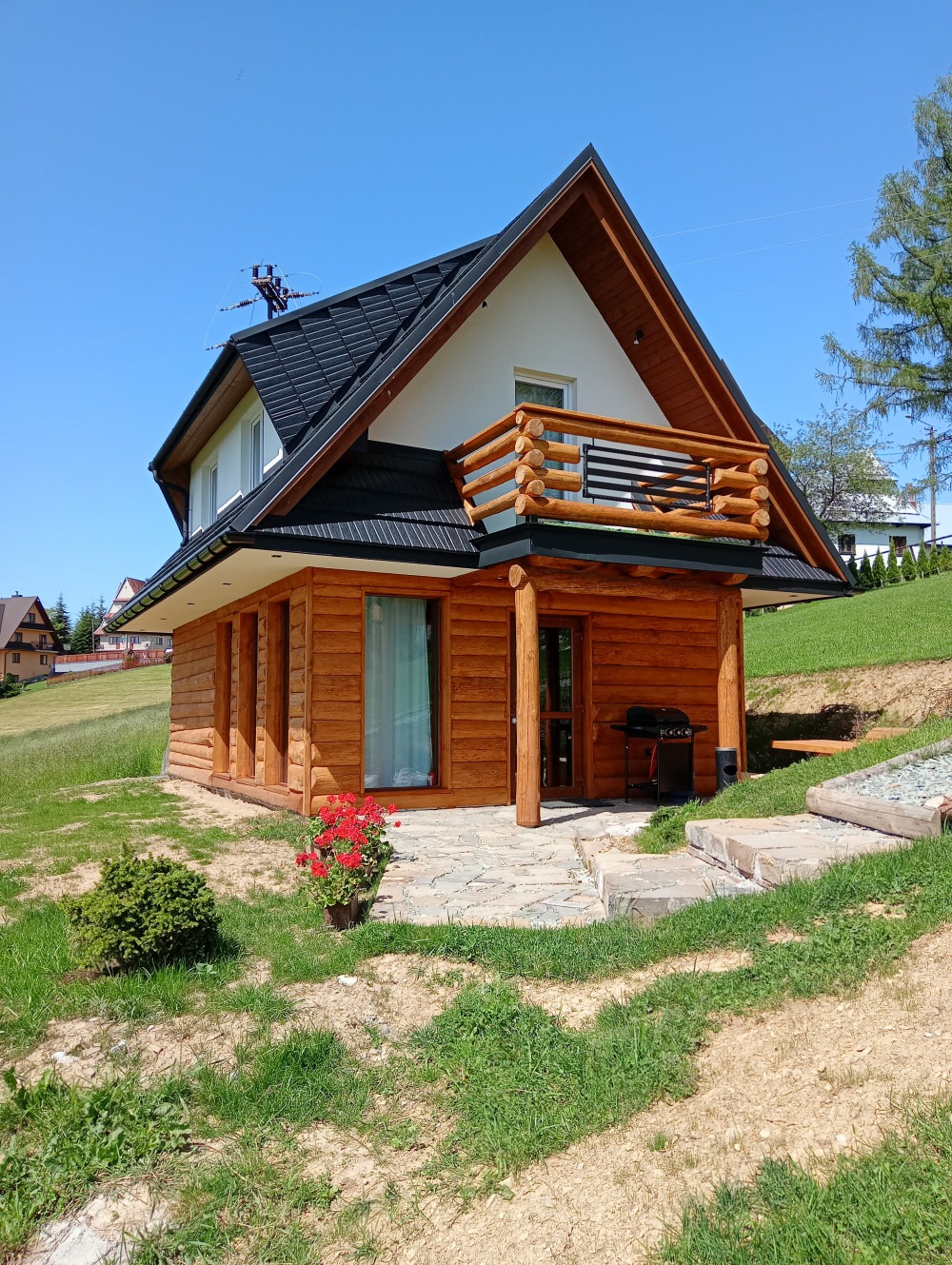 Drewniany domek z bali z balkonem, czarnym dachem i kamienną ścieżką, otoczony zielonym trawnikiem w słoneczny dzień.