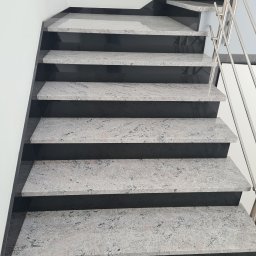 Jan Hybiak - KAMIENIARSTWO EXPORT- IMPORT - Schody wewnętrzne z szarego kamienia z ciemnymi podstopnicami i metalową balustradą, widok z dołu.