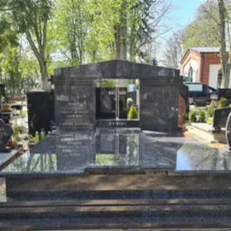 Granitowy nagrobek z wyciętym oknem na cmentarzu, z inskrypcjami i krzyżem w tle, otoczony zielenią i innymi nagrobkami w słoneczny dzień.