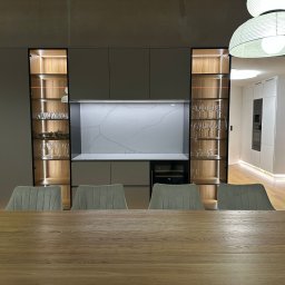 Jan Hybiak - KAMIENIARSTWO EXPORT- IMPORT - Nowoczesna kuchnia z blatem z białego kamienia z szarymi żyłami, oświetlonymi szafkami i drewnianym stołem na pierwszym planie. Widoczny fragment otwartej przestrzeni.