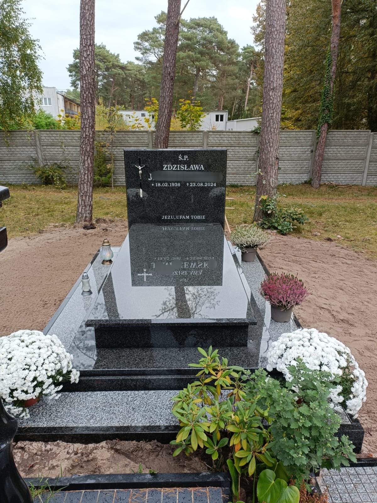 Czarny, polerowany pomnik z wygrawerowanym krzyżem i napisem 'Ś.P. Zdzisława', otoczony kwiatami w doniczkach i lampionami, na tle piaszczystego podłoża i drzew.