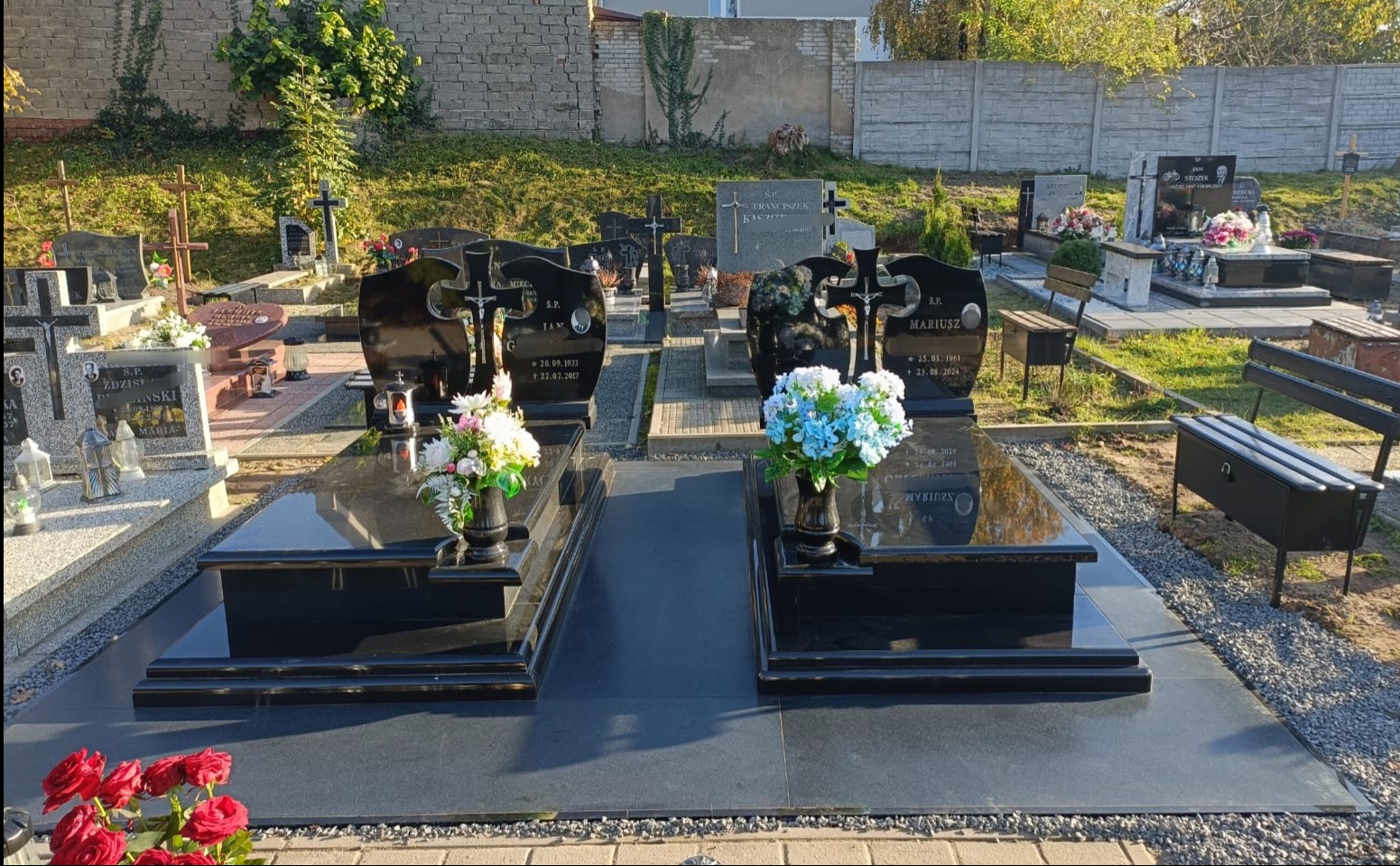 Dwa czarne, podwójne pomniki na cmentarzu z kwiatami w wazonach, otoczone żwirem i kostką brukową, w tle inne nagrobki i mur.