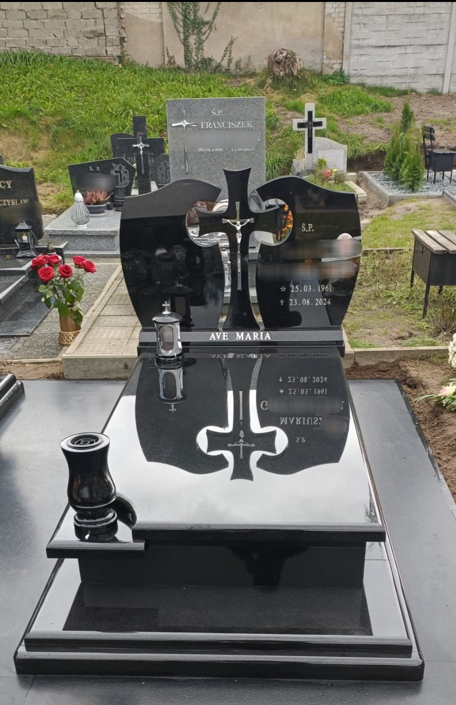 Czarny, błyszczący nagrobek z wygrawerowanym krzyżem i napisem 'Ave Maria', otoczony innymi nagrobkami na cmentarzu.