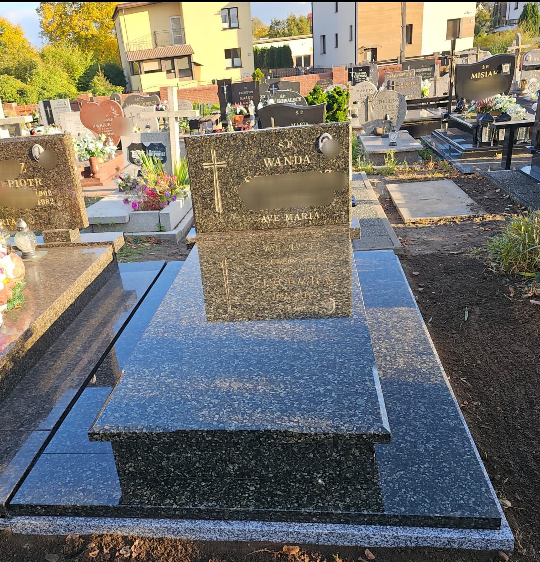 Nowoczesny nagrobek z ciemnego granitu z wygrawerowanym krzyżem i napisem, położony na cmentarzu z innymi nagrobkami w tle i budynkami w oddali.