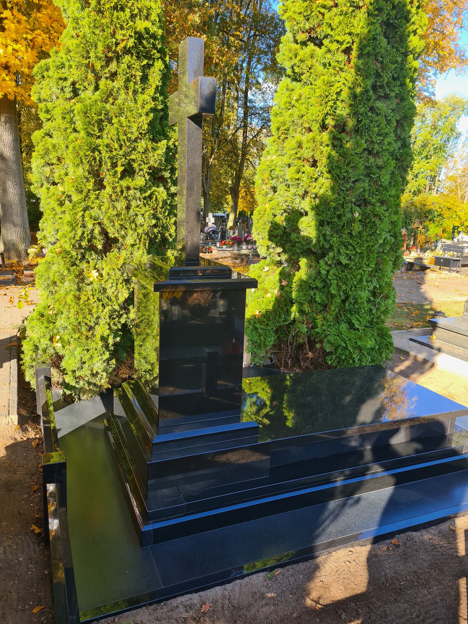 Czarny, granitowy pomnik z krzyżem, flankowany przez dwie wysokie, zielone tuje na cmentarzu w słoneczny dzień.