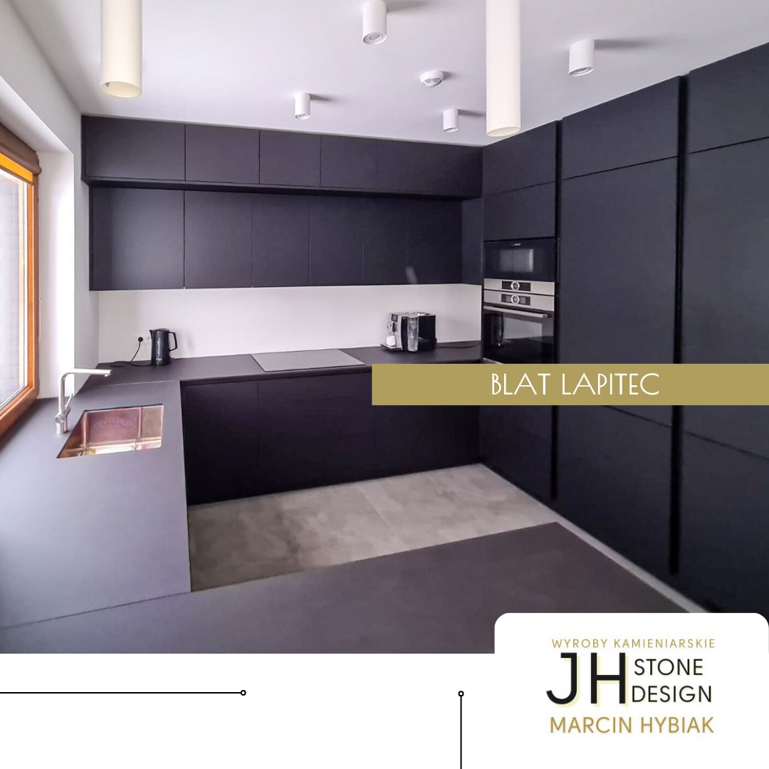 Nowoczesna kuchnia z czarnymi matowymi szafkami i blatem z ciemnego kamienia, widoczny zlewozmywak z miedzianym wykończeniem i logo firmy kamieniarskiej w rogu.