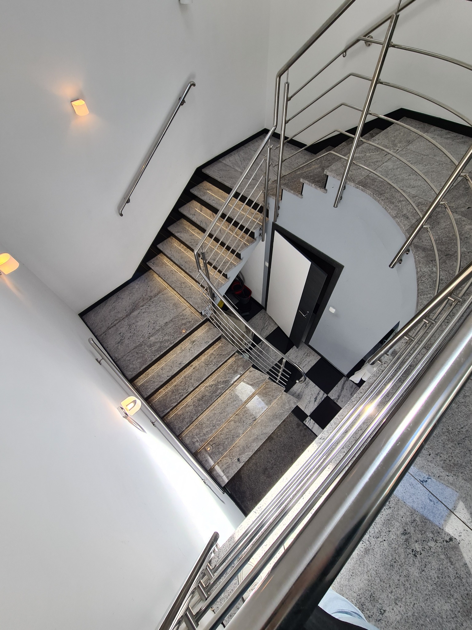 Wysoki kąt widzenia na klatkę schodową z szarymi stopniami i metalową balustradą, czarno-biała podłoga w szachownicę przy drzwiach na dole.