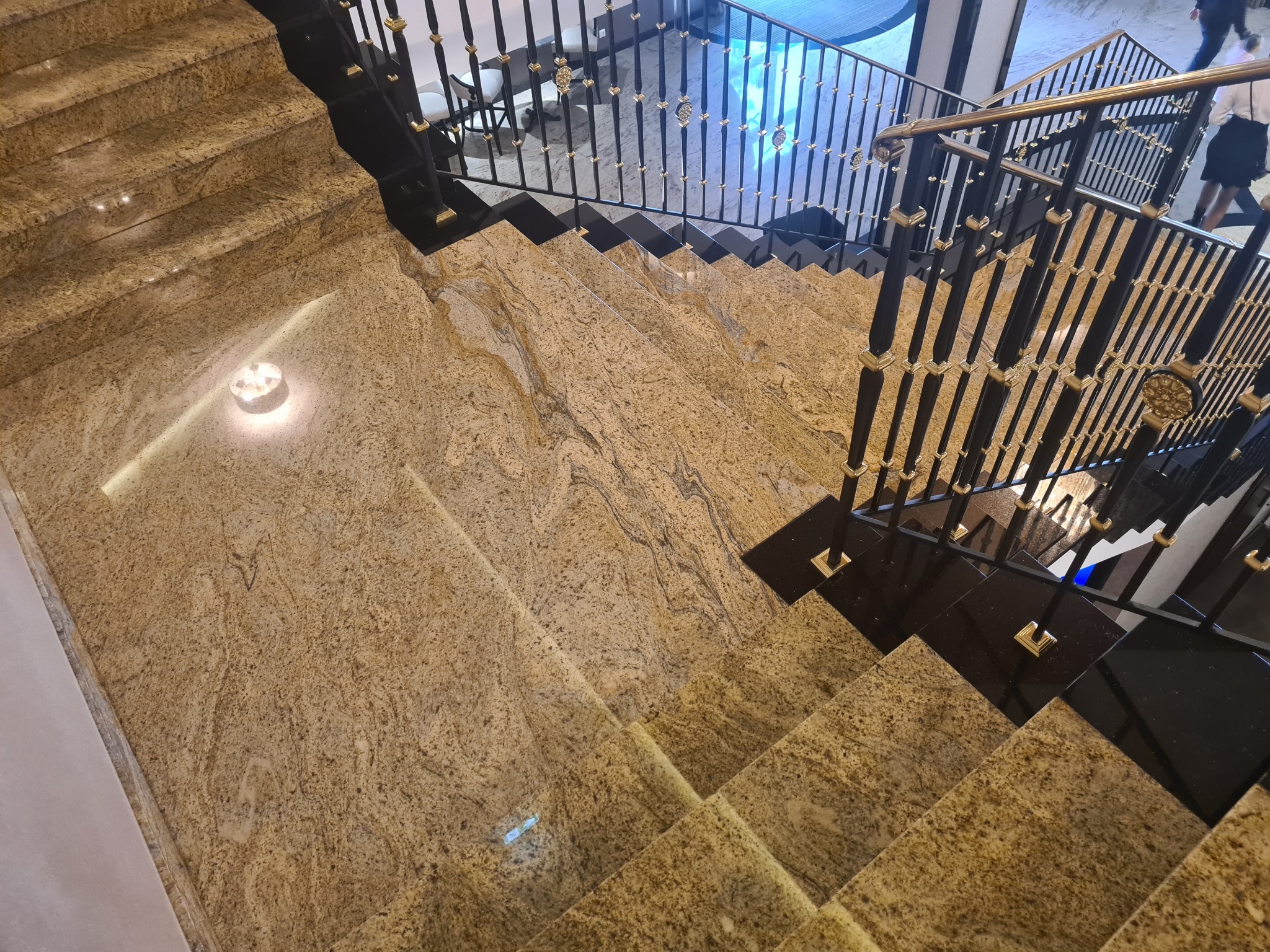 Widok z góry na eleganckie schody wewnętrzne wyłożone beżowym kamieniem z wyraźnym wzorem, z czarną balustradą z dekoracyjnymi złotymi elementami.