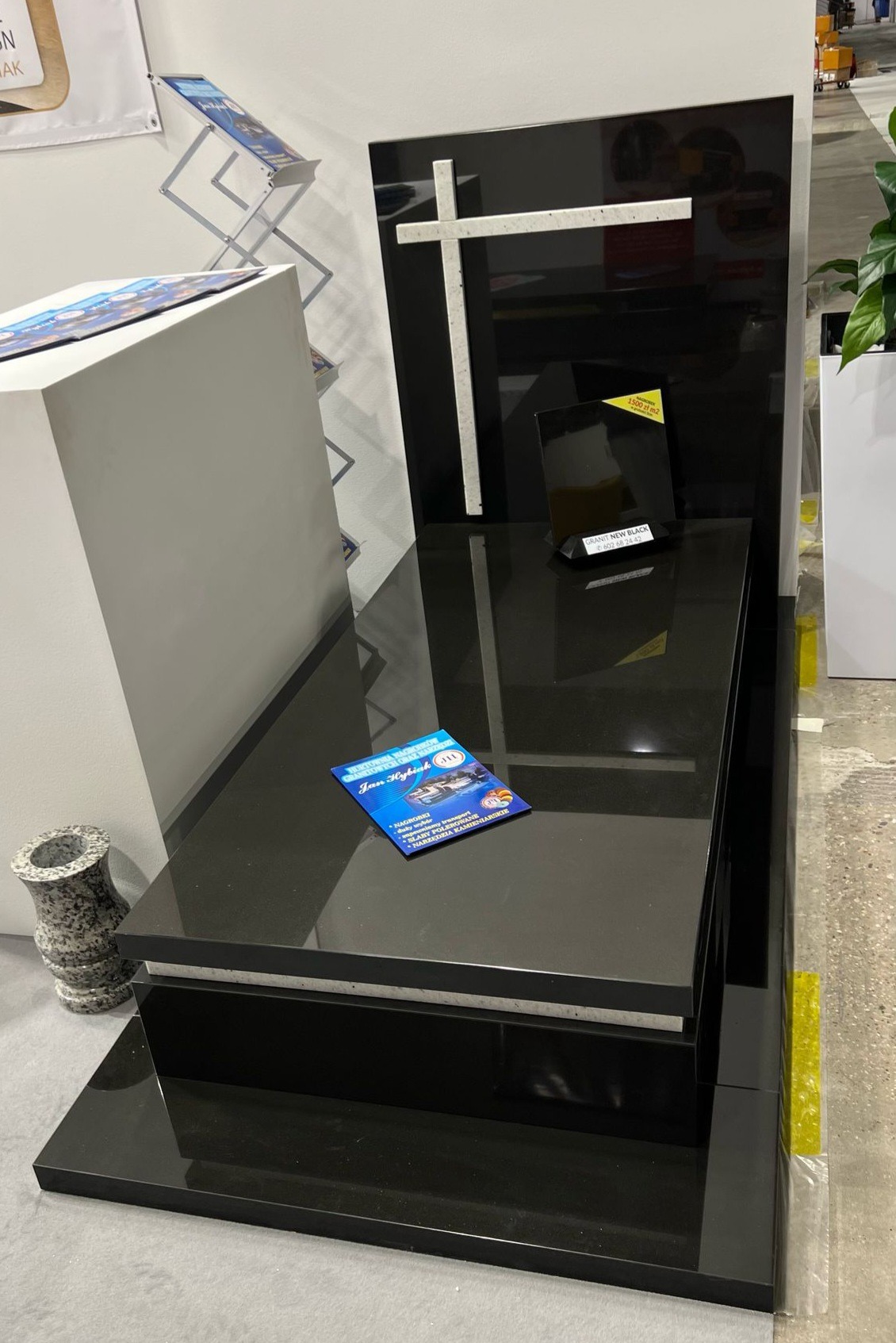 Czarny nagrobek z białym krzyżem na wystawie, ulotka informacyjna na płycie, pokazujący cenę granitu.