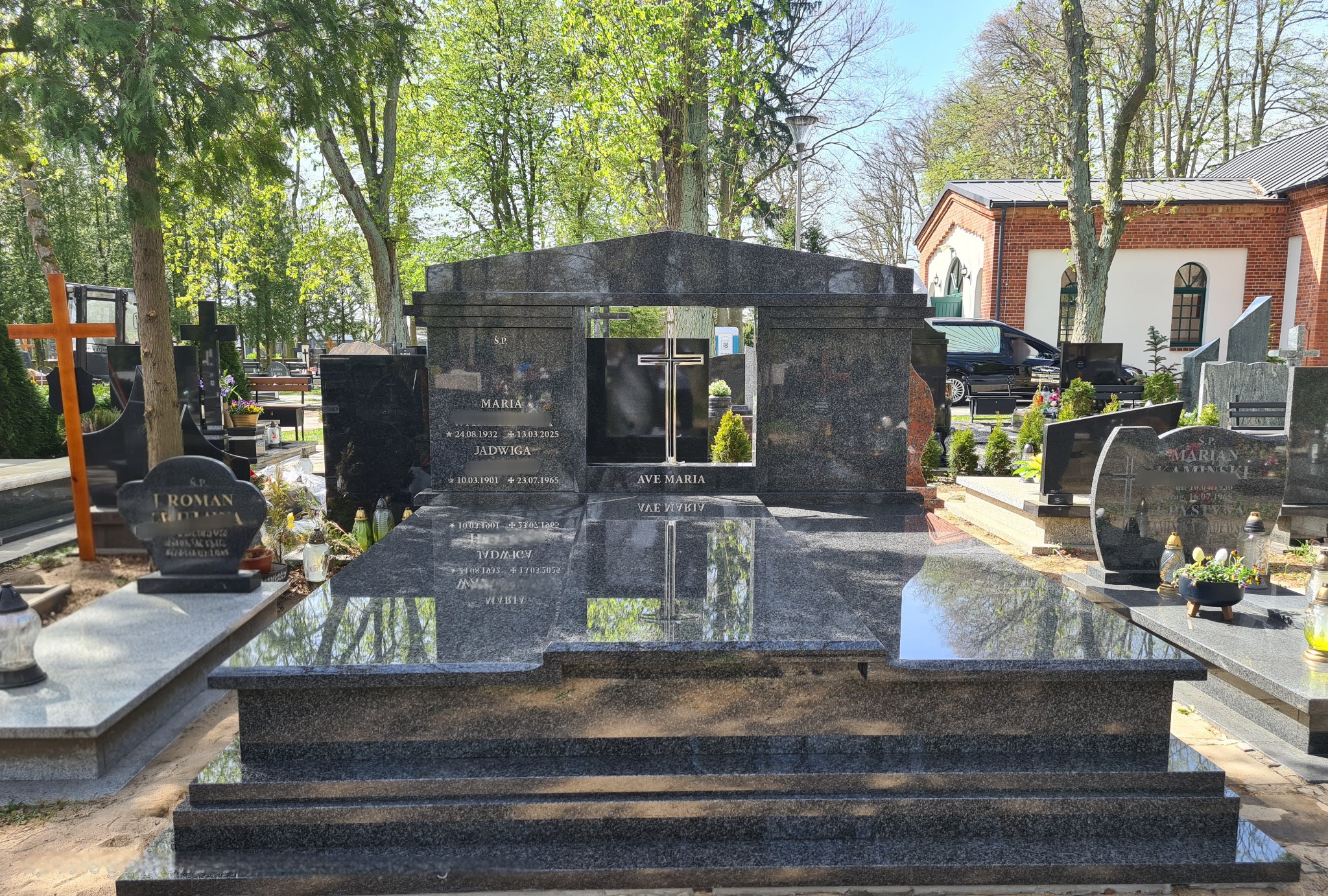 Granitowy nagrobek z wyciętym oknem na cmentarzu, z inskrypcjami i krzyżem w tle, otoczony zielenią i innymi nagrobkami w słoneczny dzień.