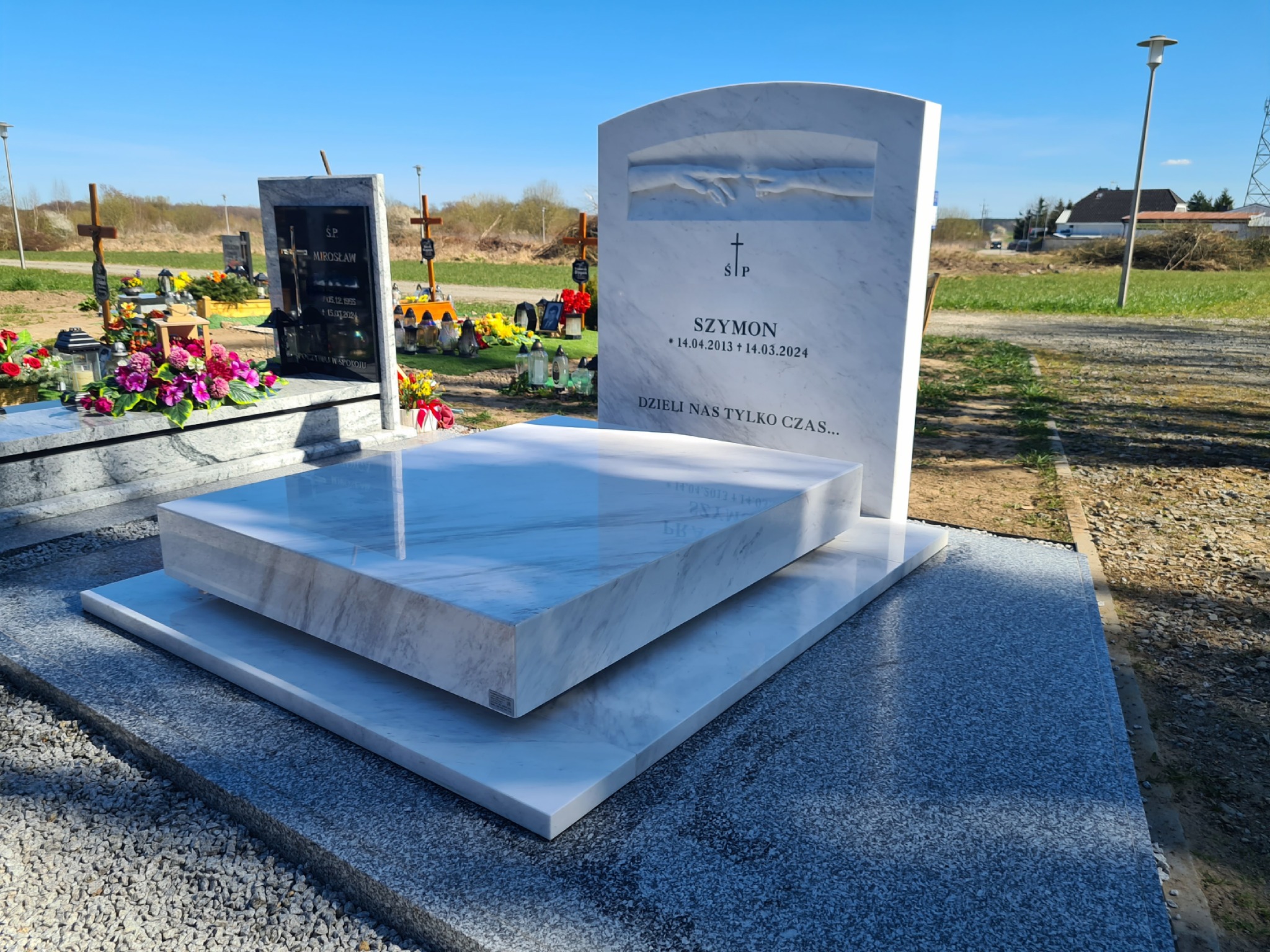 Biały marmurowy nagrobek z inskrypcją 'Szymon 14.04.2013-14.03.2024' na cmentarzu w słoneczny dzień. Rzeźbione ręce nad krzyżem.